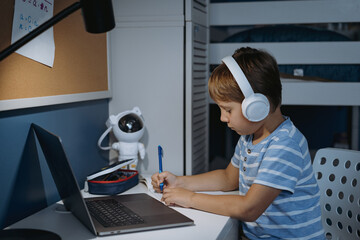 caucasian little boy wearing wireless headphones studying at home using laptop