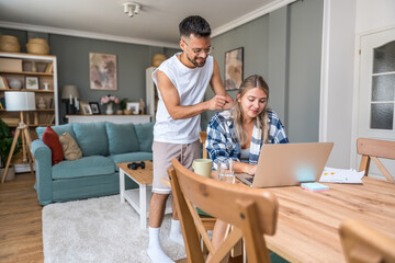 Fototapeta premium Young freelance woman working at home on laptop computer finance and economics revision while her boyfriend support her and make her company while she work Happy couple small company owners having fun