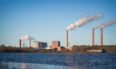 A factory with tall smokestacks emits plumes of smoke into the clear blue sky, reflecting industrial activity in a coastal area