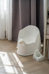 A simple, white potty chair is positioned in a bright room, bathed in soft natural light from the sheer curtains nearby