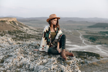 Woman in hat and sweater sitting on mountain rock edge with boots, looking out into the distance