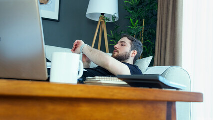 A man rest on a sofa, glancing at his watch while taking a break from work. A laptop and coffee mug rest on a wooden table. Laid back work from home routine.