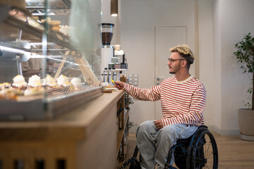 Concentrated disabled man in wheelchair paying for purchase in coffee shop using terminal, credit card. NFC technology, contactless payment, using bonus loyalty system, completing cashless transaction