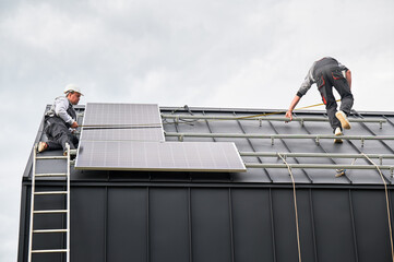 Workers measuring photovoltaic solar panels with tape measure. Men taking measurements before mounting solar modules on roof of house for generating electricity through photovoltaic effect.