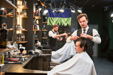 Professional barbers working on client hair in barbershop. Male hairstylists cutting hair in modern ambiance, with rustic wooden shelves stocked with various grooming products.