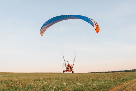 Paragliders on paraplane start to fly on red paraplane in sky above fields in countryside in summer at sunset