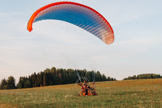 Paragliders man and woman start to fly on red paraplane in sky above fields in countryside in summer at sunset, blurred and unrecognizable