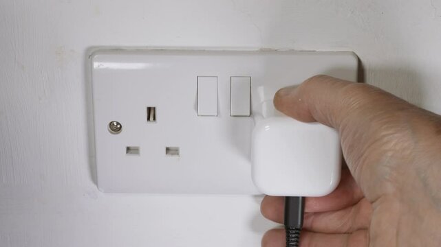 Closeup of man&rsquo;s hand switching off the power at a UK British double wall socket, then pulling out a travel plug with attached cable or cord, then pressing the three pins flat for packing.