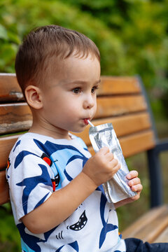 Young child enjoys a juice pouch while sitting on a park bench. A young child sips from a juice pouch, sitting contentedly on a bench in a green park.