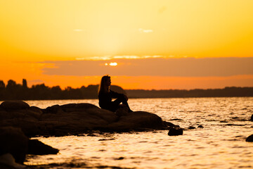 Person's silhouette on rocks at a vibrant sunset by the lake. A person sits on rocky shorelines, silhouetted against a stunning sunset over the calm lake waters.