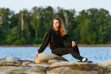 Young woman enjoying the sunset on rocky shoreline by the lake. A young woman sits on large rocks near a tranquil lake