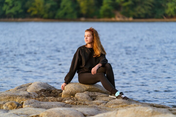 Young woman relaxing on a rock by the water at sunset. A young woman in casual attire sits on a rock by the water, enjoying the calm scenery during sunset.