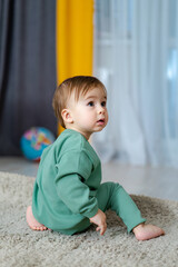 Toddler playing on the floor in a bright living room setting. A young child is sitting on a soft rug, gazing curiously at their surroundings in a cozy room filled with sunlight.