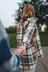 couple holding hands, young beautiful caucasian man and woman take each other hands, dressed in checkered shirts, woman leads the way in park in summer, follow me, love and family concept