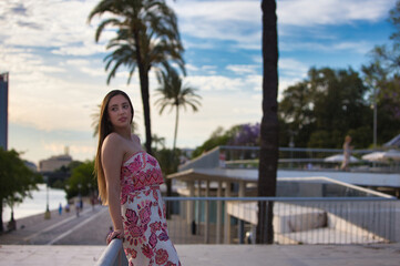 Beautiful young blonde Spanish woman resting leaning on the iron railing, in the background trees and the other bank of the river in seville. the woman is dressed in a patterned jumpsuit.