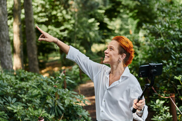 A woman with red hair smiles and films with a selfie stick in a wooded area on campus.