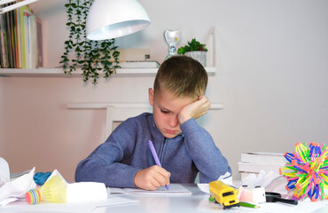 An unhappy tired schoolboy boy is doing his homework among stack of books and clutter on his desk....