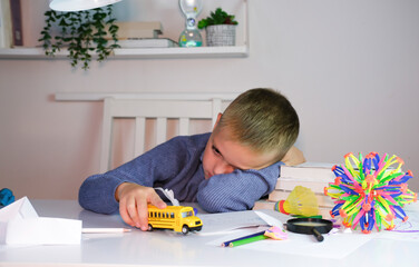 An unhappy tired schoolboy boy is doing his homework among stack of books and clutter on his desk....