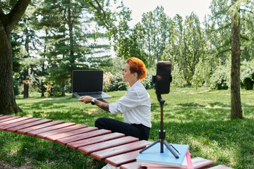Fototapeta premium A teacher records a video outdoors, sitting on a bench in a park, showcasing her laptop.