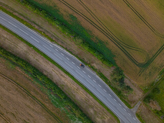 Top down photo of a solitary straight grey asphalt road cutting through a patchwork of agricultural fields. A single car is visible on the road, highlighting the tranquility of the area. Ida Virumaa.