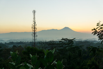 Obraz premium This image captures a peaceful morning landscape with a towering telecommunications tower set against the backdrop of a distant mountain at sunrise. 