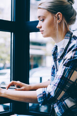 Side view of young blonde hair female in casual clothes using laptop while studying at table in modern coffee shop interior sitting alone near window. Young contemporary woman working on freelance