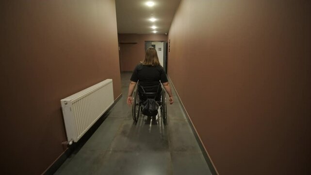 A woman in a wheelchair navigates down a long, dimly lit hallway, symbolizing independence and accessibility in a public or residential building. The scene captures determination and solitude.