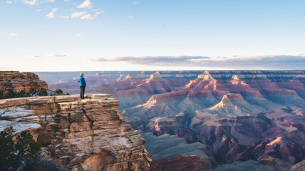 A photo of a tourist attraction, the Grand Canyon. There's a person standing on the edge of the canyon, looking out at the vast expanse below. The sky is clear, with a few clouds scattered across it. 