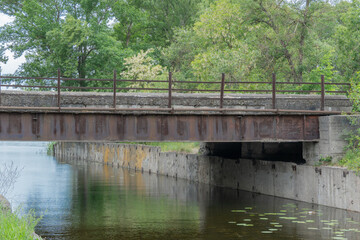 Iron concrete bridge over the river canal. Reinforced pedestrian from one bank of water to other. Picturesque trees and bushes grow along the support slabs. Empty footbridge of bridge-canal with pier.