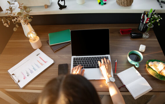 Overhead view of woman typing on laptop at tidy desk, surrounded by coffee, notebook, documents, and croissant. The organized workspace, with bright natural light, reflects productivity and focus