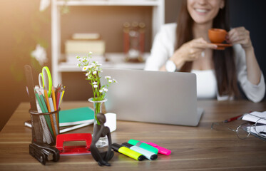Workspace featuring colorful office supplies, a laptop, and vase of flowers, with a smiling woman holding a cup of coffee in the background. The bright, organized setting exudes creativity