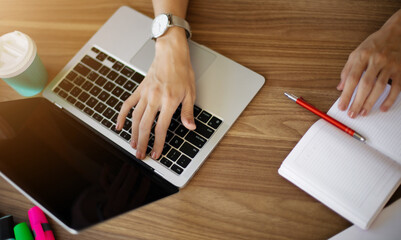A top-down view of a workspace with one hand typing on a laptop and the other writing in a...