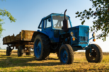 A blue, old tractor with a trailer stands near a field in the village. Morning