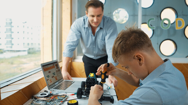 Caucasian teacher help student fixing car model and giving advise for programing system or coding prompt while highschool boy listen and use equipment to repair robot at STEM classroom. Edification.