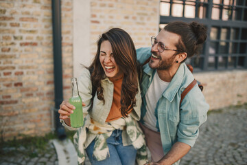 Cheerful trendy couple with bottles of drinks laughing on a street.