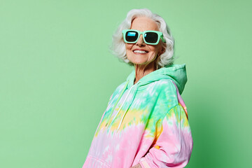 a stylish elderly woman in a tie-dye hoodie, wearing oversized sunglasses and smiling, standing against a preen background, diverse fashion model