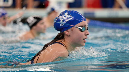 A photo of a female swimmer with a blue cap and goggles, swimming in a pool. She has long, wet hair and is wearing a swimsuit. The background is blurred and contains other swimmers and the poolside.