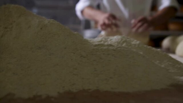 Skilled baker kneading two pieces of dough by hand on a work table, set against a background of bread-filled shelves and a stainless steel dough container. Perfect for showcasing artisan bread-making 