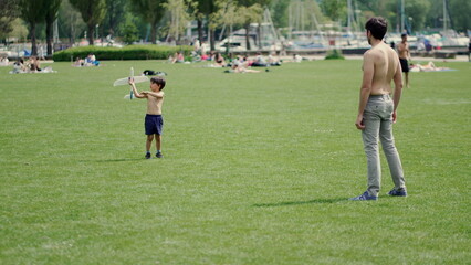 Father and son enjoy sunny day in park, launching toy airplane. Father stands watching as son prepares to launch, bonding through play and exploration amidst greenery and other families