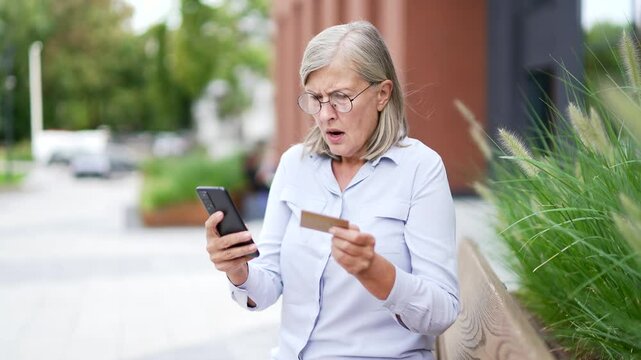 Concerned elderly woman outside holding a credit card and phone, potentially a victim of online fraud, sitting on a bench in an urban setting. Elderly female with gray hair became victim of deception