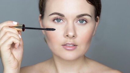 Fototapeta premium Close-up portrait of young woman on gray background, doing makeup. Mascara for eyelashes. Self care.