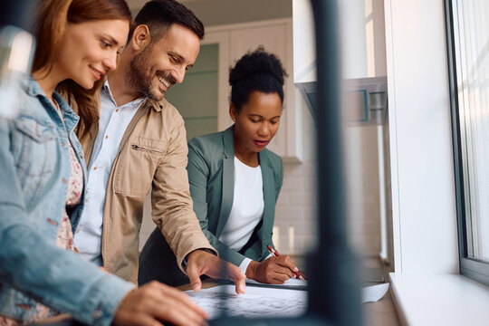Happy couple of real estate buyers analyzing blueprints with their agent in kitchen.