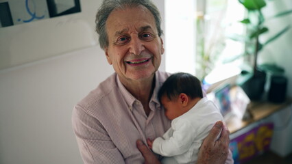 Grandfather smiling while holding newborn baby, looking directly at the camera, capturing a joyful moment of affection and bonding in a home environment