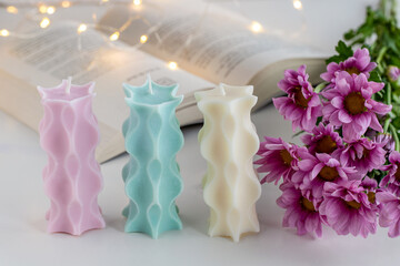 Three soy candles on a table with a book, flowers and lights in background. Home decor.
