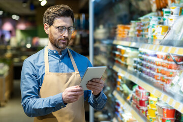 Grocery store manager conducting inventory on tablet. Supermarket setting with shelves stocked with...