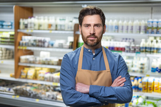Grocery store employee stands confidently in front of dairy aisle. Wearing blue shirt and apron, he demonstrates professionalism in retail setting. Background includes various products on shelves. - Powered by Adobe