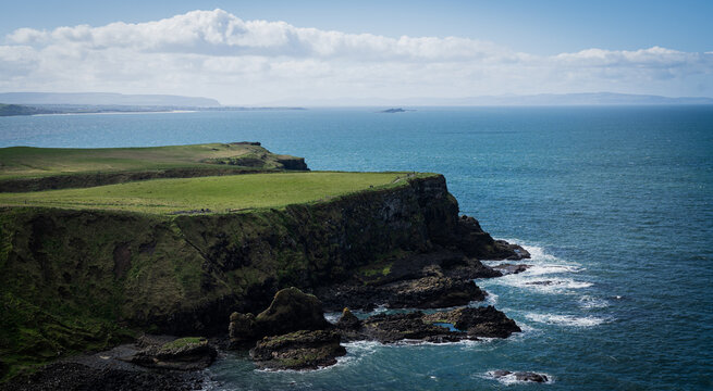 Green landscape above coastal cliffs in Ireland. The sea is blue and calm, and the sky is clear.