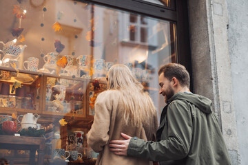 A couple admiring decorative pottery in a shop window. The woman has long hair and is wearing a beige coat, while the man in a green jacket stands close behind her, smiling.