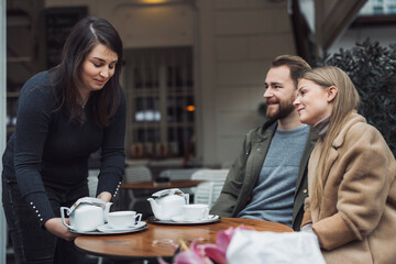 A waitress serving tea to a couple at an outdoor café. The woman is placing teapots and cups on the table while the couple looks on, smiling. The setting is cozy and inviting, with greenery in the bac