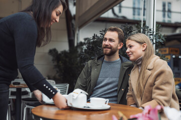 A waitress serves coffee to a couple sitting at a cozy café table. The woman, dressed in a beige coat, smiles at the man, who wears a green jacket. The café has a relaxed atmosphere with plants in the